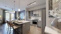This thoughtfully designed one-bedroom, one-bathroom unit offers a cohesive modern aesthetic throughout its single-floor layout. The open-concept kitchen is a standout feature, showcasing gray shaker-style cabinetry — including glass-front upper cabinets — white quartz countertops, stainless steel appliances, and a peninsula with bar seating, illuminated by pendant lights and track lighting overhead. Light wood-tone flooring runs through the kitchen and flows into the adjacent living room, where dark hardwood floors, a contrasting accent wall, and floor-to-ceiling French doors provide access to a private balcony, filling the space with natural light. The bedroom features soft carpet, a calming blue accent wall, and a large window dressed with linen drapes. It connects directly to a full bathroom with tile flooring, a soaking tub with shower, gray vanity cabinetry, and white quartz countertops. An adjoining walk-in closet offers multiple hanging rods and built-in shelving, providing generous storage. Throughout the unit, a neutral palette with modern finishes creates a clean, contemporary ambiance.