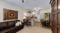 This single-floor residence showcases an open-concept layout that seamlessly connects the living, dining, and kitchen areas into one cohesive space. The living area features light carpeting, a dark leather sofa, and warm wall art, creating an inviting atmosphere anchored by a ceiling fan overhead. The kitchen is outfitted with white cabinetry, white appliances including a full-size refrigerator, and a peninsula counter with bar seating for two — ideal for casual dining or entertaining. Light tile flooring extends through the kitchen and dining areas, providing a clean contrast to the carpeted living space. The dining area accommodates a classic wooden table and chairs, accented by decorative greenery that adds a natural touch. A round decorative mirror on the wall adds visual interest to the dining zone. Ceiling fans in both the living and kitchen areas promote airflow throughout the space. The neutral color palette of whites and warm beiges throughout lends the interior a bright, airy feel, while the open floor plan maximizes the sense of space and encourages a functional, connected living experience.