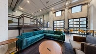 This expansive, industrial-conversion space makes a strong architectural statement through its dramatic double-height ceilings, exposed ductwork, and structural columns that reflect its warehouse origins. The interior is organized across a main floor and a raised mezzanine level connected by a steel staircase with wire-mesh railings, adding both visual interest and functional zoning to the layout.

The main floor features polished concrete underfoot and hosts multiple distinct seating areas. A generous lounge zone is anchored by a teal velvet sectional sofa paired with a round wood coffee table, offering a comfortable gathering spot. Nearby, a separate seating area includes a modular gray sectional, an accent armchair, and a wall-mounted flat-screen television, creating a more intimate setting. A row of high-top tables with yellow chairs lines the windowed perimeter, providing an informal dining or work area bathed in natural light.

The mezzanine level, overlooking the main floor, contains additional lounge seating and benefits from multiple windows framing urban views. The entry area retains raw industrial character with exposed concrete, a freight elevator, and a roll-up door. Track lighting throughout ensures well-distributed illumination across the entire space.