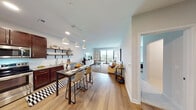 This thoughtfully designed single-floor residence offers a seamless blend of modern aesthetics and functional living. The open-concept layout connects the kitchen, dining, and living areas into one cohesive, light-filled space. Rich dark-wood cabinetry anchors the kitchen, which is outfitted with stainless steel appliances, a subway tile backsplash, light quartz countertops, and open industrial-style shelving for additional storage and display. Pendant lighting over the dining area adds a warm, contemporary touch, while light-toned hardwood flooring flows throughout the main living spaces, enhancing the sense of openness.

The living room is generously proportioned, offering room for comfortable seating arrangements and benefiting from a large sliding glass door that opens directly onto a private balcony overlooking lush tree canopy — filling the space with natural light and outdoor views.

The bedroom is a calm, carpeted retreat featuring a large window with serene views of surrounding greenery, providing both natural light and a peaceful atmosphere.

The bathroom continues the warm wood theme with dark cabinetry, a wide vanity mirror, and hardwood-style flooring, maintaining a cohesive design language across the entire residence. Overall, the space presents a clean, modern, and inviting ambiance ideal for comfortable everyday living.