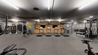 Located at 460 North Broad Street in Philadelphia, this fitness center offers a generously sized, single-floor exercise space designed to accommodate a wide range of workout styles and fitness levels. The layout follows an L-shaped floor plan, providing distinct zones for cardio, strength training, and functional fitness, all finished with durable rubber flooring throughout.

The cardio zone features a row of treadmills equipped with individual screens, positioned in front of a warm wood-toned built-in shelving unit with mounted flat-screen TVs — a motivating focal point anchored by a bold "You Got This" wall graphic. Ellipticals and rowing machines round out the cardio offerings.

The strength training section is well-appointed with a cable machine system, a dedicated free weights area stocked with a full dumbbell rack, barbells, weight plates, squat racks, and multiple adjustable benches. Additional selectorized machines line the perimeter for targeted muscle group training.

A separate functional training area provides open floor space for dynamic workouts, complete with plyo boxes, sandbags, a heavy bag, and resistance tools. Large floor-to-ceiling mirrors expand the sense of space throughout.

The industrial-modern aesthetic is defined by painted brick walls in cool gray tones, exposed ceiling infrastructure, and recessed and strip lighting that keeps the environment bright and energized. Built-in wooden cubbies offer convenient storage for personal belongings.