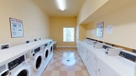 This shared laundry facility is designed with practicality and efficiency in mind, offering a clean and organized environment for residents. The room features a row of commercial-grade front-loading washing machines along one wall, complemented by a full lineup of top-loading dryers along the opposite side, providing ample capacity to accommodate multiple users simultaneously. The space is finished with warm yellow walls that create a welcoming atmosphere, while the classic checkered tile flooring in soft pink and white tones adds a touch of character underfoot. Overhead fluorescent lighting ensures the room is well-lit throughout the day, and a centrally positioned window along the back wall allows natural light to filter in, offering a view of the surrounding greenery. The vaulted ceiling height adds a sense of openness to the space. Instructional signage is posted on the walls to assist users with proper machine operation. A utility shelf is conveniently positioned near the window, providing a surface for folding or sorting laundry. The facility is thoughtfully laid out to maximize usability, making it a fully functional and comfortable communal laundry space.