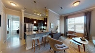 This well-appointed single-floor residence offers a thoughtfully designed open-concept layout that seamlessly connects the kitchen, dining, and living areas. The kitchen is a standout feature, showcasing rich dark cabinetry, granite countertops, stainless steel appliances including a refrigerator, range, and built-in microwave, and a breakfast bar with bar stools — all accented by elegant pendant lighting and a track light fixture overhead. A tile backsplash adds a clean, polished touch. Adjacent to the kitchen, the dining area accommodates a round wood table with modern seating, set beneath tasteful wall art that adds character to the space. The living room is bright and welcoming, with large windows dressed in layered blinds and drapes that allow generous natural light. Crown molding runs throughout, lending an elevated finish to the neutral-toned walls and wood-look flooring. Both bedrooms are comfortably sized with ample natural light, large windows, and warm, contemporary décor. The bathroom features granite countertops, dark wood cabinetry, and a full soaking tub with tiled surround. A hallway connects all areas of the residence, creating a cohesive and functional flow throughout this polished, modern living space.