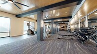 This expansive fitness center is designed with a contemporary aesthetic, combining industrial and modern elements throughout its single-level layout. The space is defined by a patterned, dark gray carpet with subtle light veining that runs the length of the floor, complementing the warm wood-paneled ceiling punctuated by bold dark structural beams and ceiling fans that enhance air circulation across the open floor plan.

The cardio zone lines one side of the facility, featuring a row of Matrix treadmills, stair climbers, stationary bikes, and elliptical machines positioned alongside large windows that flood the area with natural light and offer outward views. On the opposing side, a dedicated strength training zone houses Matrix cable machines, power racks, barbells, and a full dumbbell rack, all set against a wall of floor-to-ceiling mirrors framed in dark trim with vertical sconce lighting. An exposed brick accent wall serves as a striking architectural focal point, adding texture and warmth to the interior.

Recessed lighting throughout ensures consistent illumination, while a separate room accessible through a brick-framed opening appears to provide additional amenity space. The overall ambiance is polished, well-equipped, and purposefully designed for a comprehensive fitness experience.