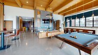 Located at 1005 Gramsie Road in Shoreview, Minnesota, this expansive single-floor amenity space is designed to impress with its thoughtful blend of industrial and contemporary design elements. The grand entrance lobby greets visitors with soaring double-height ceilings, floor-to-ceiling glass doors, warm wood beam accents, teal accent walls, and a sculptural chandelier, setting a welcoming and sophisticated tone throughout.

Polished concrete floors extend across the entire space, reflecting natural light that pours in through large glazed walls and sliding glass doors that open directly to an outdoor patio and courtyard. The open-plan layout flows seamlessly between distinct functional zones. A staffed reception desk with a sleek metal fascia anchors the lobby, while a lounge area nearby features rich leather seating and lush greenery.

Deeper into the space, a dramatic lounge zone centers around a linear fireplace set beneath a textured, sculptural tile feature wall, surrounded by leather chairs and a generous sectional sofa. Exposed ductwork and structural columns add an industrial character. An adjacent dining and bar area offers booth-style yellow seating against a patterned accent wall beneath a coffered wood-beam ceiling. A games area completes the space, equipped with a billiards table and shuffleboard, alongside artwork-adorned concrete walls that lend an urban gallery feel.