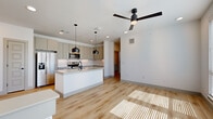 This contemporary two-story residence offers a thoughtfully designed layout spread across two levels. The main floor is anchored by a bright, open-concept living and kitchen area bathed in natural light streaming through large windows with horizontal blinds. The modern kitchen is a standout feature, showcasing sleek grey cabinetry, a white tile backsplash, stainless steel appliances including a French door refrigerator and electric range, and a generous center island topped with granite counters and a matte black sink fixture. Stylish black pendant lights hang above the island, complemented by recessed lighting and a ceiling fan overhead, creating a well-lit and functional space. Light hardwood-style flooring runs throughout the main living areas, adding warmth to the clean, neutral palette. The bedroom is a comfortable retreat featuring plush carpeting, a ceiling fan, and natural light from a pair of windows. It connects directly to the full bathroom via a sleek sliding barn door, lending the space a modern, architectural touch. The bathroom is well-appointed with grey cabinetry, a granite vanity, an LED-backlit mirror with matte black fixtures, and a tiled tub-shower combination. A dedicated closet and staircase complete the functional layout of this well-finished home.