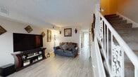 This two-story home offers a thoughtfully arranged layout across its two levels, combining practical design with a warm, lived-in aesthetic. The main living area serves as the heart of the home, featuring light wood-look flooring, a textured ceiling, and neutral walls that create a bright, open atmosphere. Wall-mounted sconces and a floor lamp provide ambient lighting, complementing the large flat-screen television set atop a dark wood media console decorated with small plants and ornamental accents. Leather sofas with decorative pillows anchor the seating arrangement, while framed artwork and diamond-shaped wall décor add visual interest throughout. A white-railed staircase connects the two floors, positioned along one side of the living space.

The kitchen is efficiently designed with white cabinetry, dark granite-style countertops, and light wood-look tile flooring. It is equipped with a full-size refrigerator, microwave, and range. Adjacent to the kitchen, a dining area benefits from natural light streaming through vertical-blind-covered windows, with a dark wood dining table and matching chairs providing a functional gathering space. A carpeted bedroom offers a cozy retreat, while a clean, simply appointed bathroom completes the home's interior.