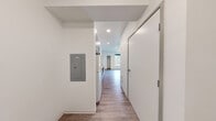 This studio unit at Koz on 317 in Tacoma, Washington offers a smartly designed, single-floor layout with one bedroom and one bathroom. Wood-look plank flooring runs throughout the entire unit, providing a cohesive and contemporary feel. The entry hallway leads past a built-in electrical panel and into the main living area, where recessed lighting and clean white walls create a bright, open atmosphere. The kitchenette is efficiently arranged along one wall, featuring white upper cabinetry, gray lower cabinets, open shelving, a stainless steel sink, a built-in microwave, and a full-size refrigerator — offering practical functionality within a compact footprint. The bedroom area is well-proportioned and benefits from a large window that frames natural tree-lined views, a ceiling fan with light fixture, and a sliding-door closet with built-in shelving for storage. A thermostat is conveniently located on the bedroom wall. The bathroom is finished with consistent wood-look flooring, a white vanity with modern hardware, a toilet, a towel bar, and a walk-in shower stall. The unit's minimalist design, neutral palette, and efficient use of space make it a functional and modern living option in Tacoma.