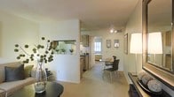 This single-floor residence is designed with an open, flowing layout that connects the living, dining, and kitchen areas into one cohesive space. A defining architectural feature is the half-wall with a pass-through window that separates the kitchen from the living area, allowing natural sightlines throughout while maintaining a subtle sense of division between spaces.

The kitchen is equipped with wood-toned cabinetry and a dark countertop, offering a warm contrast against the light-colored walls and flooring. The pass-through ledge doubles as a convenient serving area, enhancing the functional design. The dining area is positioned centrally, furnished with a round table and modern chairs beneath a ceiling-mounted light fixture, creating an intimate yet accessible dining environment.

The living space features neutral carpeting and smooth walls in a soft, warm tone, contributing to a bright and welcoming ambiance. Wall-mounted framed art and decorative lighting fixtures add subtle character to the space. A glimpse through the hallway reveals a naturally lit room beyond, suggesting well-distributed light throughout the residence. Overall, the layout prioritizes practicality and openness.