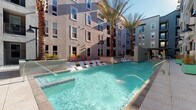 This expansive outdoor amenity courtyard is designed to deliver a resort-like experience with a diverse array of recreational and social features. At its centerpiece is a shimmering resort-style swimming pool flanked by sleek white lounge chairs, complemented by a separate spa with bubbling jets — all framed by tall palm trees and the multi-story building facade. String lights overhead add a warm ambiance that transitions seamlessly from day to evening.

The fully equipped outdoor kitchen is a standout feature, showcasing multiple stainless steel grills set into concrete countertops, a utility sink, and ample prep space beneath a retractable pergola — ideal for al fresco dining and entertaining. Adjacent to the kitchen, a fire pit lounge area with a concrete gas fire table is surrounded by woven lounge chairs and bar-height seating, creating an inviting gathering spot.

For recreation, the courtyard includes a dedicated bocce ball court shaded by a bold orange canopy pergola, as well as a concrete ping pong table set on a turf surface. Planter beds with ornamental grasses, river rock landscaping, and carefully placed greenery add natural texture throughout the space, while the open-sky design ensures abundant natural light across the entire courtyard.