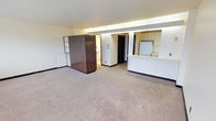 This single-floor space offers an open, functional layout designed to maximize usability. The main living area is finished with neutral wall-to-wall carpeting that lends a clean, understated look throughout. A distinctive dark wood-paneled wardrobe or room divider anchors one side of the space, providing both storage and a visual focal point that contrasts with the light-toned walls and ceiling.

Toward the back, a kitchen area is integrated into the floor plan, featuring white upper cabinetry, a countertop bar overhang suitable for casual seating, and a full-size refrigerator. The kitchen is separated from the main living space by a low partition, creating a defined zone while maintaining an open feel.

Adjacent to the kitchen, a doorway leads to a secondary area with tiled flooring, suggesting a bathroom or utility space. A dark interior door nearby offers access to an additional room, providing further functional separation within the single-floor layout.

Recessed lighting runs along the upper perimeter of the space, casting an even, ambient glow that enhances the room's clean aesthetic. The textured ceiling and simple trim work contribute to a practical, no-frills design. Overall, the space is thoughtfully arranged to balance open living with organized, defined functional areas.