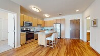 This single-floor residence is thoughtfully designed with a modern, functional layout connecting its key living spaces through warm hardwood flooring that runs throughout. The kitchen serves as a central hub, outfitted with light maple cabinetry extending to the ceiling for ample storage, granite countertops, and a full suite of stainless steel appliances including a gas range, microwave, dishwasher, and refrigerator. A freestanding kitchen island with a granite surface and seating for two adds both workspace and casual dining functionality.

The living area is bright and welcoming, benefiting from floor-to-ceiling windows and a glass door that opens directly to a private balcony with urban views. This generous space is well-suited for both relaxing and entertaining.

The bedrooms are carpeted and comfortably sized, each featuring windows that bring in natural light and views of the surrounding cityscape. The bathroom includes a full tub-shower combination with white tile surround, a wood-toned vanity, and hardwood flooring. A dedicated laundry area houses a full-size top-loading washer and dryer with overhead wire shelving for additional storage, completing this well-appointed residence.