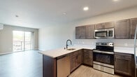 This well-appointed single-floor residence showcases a thoughtfully designed open-concept layout that seamlessly connects the kitchen and living areas. The kitchen is a standout feature, equipped with dark espresso cabinetry, crisp white quartz countertops, and a full suite of stainless steel appliances including a range, microwave, dishwasher, and refrigerator. A peninsula with an undermount sink and matte black faucet provides additional prep space and a natural division between the kitchen and the living area. Light wood-tone flooring runs throughout the main living spaces, complementing the neutral wall tones and recessed lighting overhead. Sliding glass doors lead to a private balcony, allowing natural light to flow into the open living area. The bedroom is comfortably carpeted and includes a ceiling fan with light fixture, a wall-mounted mini-split climate control unit, and a window with exterior views. The bathroom is finished with large-format tile in the walk-in shower with a sleek black-framed glass door, a wall-hung toilet, a vanity with white countertop, dark cabinetry, and an illuminated mirror. A stacked washer and dryer unit is conveniently integrated within the unit, adding practical in-home laundry access.