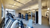 This fitness center offers a comprehensive workout environment designed to accommodate a variety of exercise routines. The space is generously sized, with an open floor plan that allows for easy navigation between equipment stations. Overhead, an exposed ceiling with industrial-style linear lighting fixtures provides bright, even illumination throughout the room, contributing to an energetic and functional atmosphere.

The cardio section is well-stocked with treadmills, ellipticals, and recumbent bikes, positioned to allow users ample personal space during workouts. Toward the center and rear of the facility, a robust selection of cable and resistance machines supports a full range of strength-training exercises. Along the far wall, a dedicated free weights area includes racks and benches, while stability balls and additional accessories are neatly arranged nearby.

The flooring features a combination of carpet and rubber mat sections, providing appropriate surfaces for both machine use and functional training. Large windows along one wall invite natural light into the space, enhancing the overall ambiance. The facility's layout and equipment selection reflect a thoughtful design focused on usability, safety, and versatility, making it suitable for users of varying fitness levels and training goals.