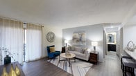 This single-floor residence offers a thoughtfully designed living space that combines modern aesthetics with everyday comfort. The living room serves as the heart of the home, showcasing smooth, wood-style flooring that adds warmth and visual continuity throughout the space. A textured ceiling adds subtle architectural interest overhead, while soft, neutral-toned walls — accented by a calming blue-gray feature wall — create a serene and inviting atmosphere. Natural light filters generously into the room through a large window dressed with sheer linen-style curtains, brightening the space throughout the day. The layout comfortably accommodates a classic sofa paired with a bold blue accent chair, a round coffee table, and coordinating side tables, all anchored by a geometric area rug that ties the design together. A curated piece of abstract artwork adorns the feature wall, complementing the space's refined yet approachable style. The overall ambiance is clean, modern, and welcoming — ideal for both relaxed everyday living and casual entertaining. The single-floor layout ensures seamless flow and accessibility throughout the home.
