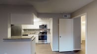 This single-floor unit is designed with a clean, functional layout that flows naturally from one area to the next. The living area showcases dark hardwood-style flooring and neutral grey walls, creating a modern, understated aesthetic. A ceiling fan with integrated lighting provides both ventilation and ambient illumination, while a large window fitted with horizontal blinds allows for natural light and privacy control. The entry door opens directly into this open living space.

The kitchen is efficiently arranged and opens to the living area via a breakfast bar with granite-style countertops, offering a seamless connection between the two spaces. White cabinetry provides ample storage, while stainless steel appliances — including a gas range, over-the-range microwave, and refrigerator — contribute a polished, contemporary look. Light wood-tone flooring in the kitchen contrasts subtly with the darker flooring in the living space.

The bedroom mirrors the living area's aesthetic, featuring the same dark hardwood-style flooring, neutral walls, a ceiling fan, and a window that brings in natural light.

The bathroom is finished with clean white subway tile lining the walls of the shower and tub combination, complemented by chrome fixtures for a simple, classic look throughout.