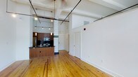 This single-floor loft showcases a compelling blend of industrial character and modern refinement. The open-concept layout is anchored by warm, light-toned hardwood floors that run throughout the space, reflecting natural light streaming in through large windows dressed with horizontal blinds. Soaring ceilings with exposed structural beams, metal conduit runs, and pendant lighting reinforce the loft's industrial heritage while maintaining a bright, airy atmosphere.

A standout architectural detail is the painted cinder block column near the entry, which adds raw, textural contrast to the otherwise clean white walls. The column features a distinctive dark green metal cap, lending a unique, historic character to the space.

The kitchen is thoughtfully designed with rich wood cabinetry, dark countertops, stainless steel appliances including a microwave and range, and a peninsula that opens to the main living area — ideal for both daily living and entertaining. Track lighting and pendant fixtures above the kitchen provide focused illumination throughout.

A black-framed glass entry door adds a sleek, urban touch and allows additional natural light into the interior. The overall ambiance is open, inviting, and full of character — a space that successfully honors its industrial roots while offering a refined, contemporary living environment.