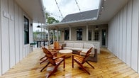 Located at 1235 Ashley Gardens Blvd in Charleston, SC, this single-level amenity pavilion is thoughtfully designed to serve as a communal gathering hub. The structure is clad in classic board-and-batten siding in a neutral palette, complementing the surrounding residential community and mature tree canopy.

The centerpiece of the pavilion is an expansive open-air deck finished in natural pine hardwood planking, which extends generously to accommodate multiple seating areas. Festoon string lights crisscross overhead, creating an inviting atmosphere for both daytime and evening gatherings. The lounge area is furnished with Adirondack chairs, a wicker sectional sofa with neutral cushions, and a round accent table, all arranged to encourage relaxed social interaction.

An integrated outdoor kitchen station anchors one side of the deck, featuring a granite countertop and stainless steel appliances, making the space well-suited for catering and community events. Pendant barn lights mounted throughout provide consistent lighting across the covered sections of the deck.

The pavilion also includes an enclosed interior mailroom, lined wall-to-wall with individual resident mailboxes and package lockers set against white wainscoted walls and the same warm pine flooring. A covered entry with a black metal handrail provides accessible access to the structure, with views of the landscaped parking and community grounds beyond.