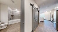 Located at 2300 Laurel Rd E in Nokomis, Florida, this single-floor residence offers a clean, contemporary design with thoughtful finishes throughout. The open-concept layout centers around a modern kitchen featuring crisp white shaker cabinetry, a subway tile backsplash, stainless steel appliances including a refrigerator, range, and microwave, and a central island with quartz countertops and an undermount stainless steel sink. Pendant globe lights hang above the island, adding a stylish focal point, while large windows dressed with white blinds fill the living area with natural light. Light-toned wood-look flooring runs consistently throughout the space, lending warmth and visual continuity. A distinctive hallway features dual sliding barn doors with matte black hardware, offering a modern industrial accent while providing practical space-saving functionality. One door leads to a well-designed bathroom complete with white shaker vanity, quartz countertop, a large frameless mirror, and a tiled tub-and-shower combination with a curved glass door. The neutral color palette of whites, soft grays, and warm wood tones creates a cohesive, inviting atmosphere suitable for a variety of interior styles.