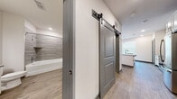 Located at 2300 Laurel Rd E in Nokomis, Florida, this single-floor residence offers a clean, contemporary design with thoughtful finishes throughout. The open-concept layout centers around a modern kitchen featuring crisp white shaker cabinetry, a subway tile backsplash, stainless steel appliances including a refrigerator, range, and microwave, and a central island with quartz countertops and an undermount stainless steel sink. Pendant globe lights hang above the island, adding a stylish focal point, while large windows dressed with white blinds fill the living area with natural light. Light-toned wood-look flooring runs consistently throughout the space, lending warmth and visual continuity. A distinctive hallway features dual sliding barn doors with matte black hardware, offering a modern industrial accent while providing practical space-saving functionality. One door leads to a well-designed bathroom complete with white shaker vanity, quartz countertop, a large frameless mirror, and a tiled tub-and-shower combination with a curved glass door. The neutral color palette of whites, soft grays, and warm wood tones creates a cohesive, inviting atmosphere suitable for a variety of interior styles.