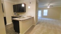 This single-floor residence offers a thoughtfully designed open-concept layout that seamlessly connects the kitchen and living areas. The kitchen is appointed with rich dark cabinetry, granite-style countertops, and a built-in sink set into a breakfast bar peninsula — an ideal spot for casual dining or entertaining. Two pendant lights hang above the peninsula, providing focused task lighting and a stylish focal point. A classic tile backsplash complements the dark cabinetry, adding texture and depth to the space. Crown molding runs throughout the ceiling, lending a refined architectural detail to the interior. The flooring transitions from tile in the kitchen to plush wall-to-wall carpeting in the living area, creating a warm and comfortable atmosphere. The living area is generously proportioned, illuminated by a ceiling fan with integrated lighting and benefiting from abundant natural light streaming through a set of bright windows and a glass-paneled door that leads to the exterior. Neutral, warm-toned walls throughout create a cohesive and inviting ambiance, offering a versatile backdrop for a variety of interior styles. The overall design balances practicality with comfort, making this a functional and welcoming living space.