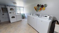 This shared laundry facility is designed with both functionality and comfort in mind. The space is outfitted with multiple top-loading washing machines and stacked washer-dryer units, providing ample capacity to accommodate residents' laundry needs efficiently. The machines are arranged to allow easy access and comfortable movement throughout the room.

The flooring consists of neutral stone-tiled surfaces that are durable and easy to maintain, contributing to the overall clean and tidy atmosphere of the space. A large window on one wall allows natural light to fill the room, creating a bright and pleasant environment. A small potted plant near the window adds a touch of greenery and warmth to the utilitarian setting.

The walls are finished in a soft, light gray tone that keeps the space feeling open and fresh. A decorative wall arrangement of colorful ceramic disc art adds a cheerful, artistic element to the room, elevating the ambiance beyond a standard utility space. The overall design strikes a practical yet inviting balance, ensuring that residents have access to a well-maintained, aesthetically considerate laundry facility.
