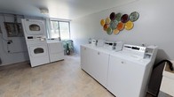This shared laundry facility is designed with both functionality and comfort in mind. The space is outfitted with multiple top-loading washing machines and stacked washer-dryer units, providing ample capacity to accommodate residents' laundry needs efficiently. The machines are arranged to allow easy access and comfortable movement throughout the room.

The flooring consists of neutral stone-tiled surfaces that are durable and easy to maintain, contributing to the overall clean and tidy atmosphere of the space. A large window on one wall allows natural light to fill the room, creating a bright and pleasant environment. A small potted plant near the window adds a touch of greenery and warmth to the utilitarian setting.

The walls are finished in a soft, light gray tone that keeps the space feeling open and fresh. A decorative wall arrangement of colorful ceramic disc art adds a cheerful, artistic element to the room, elevating the ambiance beyond a standard utility space. The overall design strikes a practical yet inviting balance, ensuring that residents have access to a well-maintained, aesthetically considerate laundry facility.