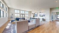 This single-floor amenity space is designed with a modern, open-concept layout that seamlessly blends comfort and functionality. The lounge area is anchored by warm wood-look flooring and large area rugs, furnished with contemporary modular seating arranged to encourage both relaxed gatherings and casual conversation. Towering windows line multiple walls, flooding the interior with abundant natural light and offering views of an outdoor pool and surrounding landscape. Glass doors provide direct access to the outdoor terrace.

The bar and kitchen area features a striking design contrast, with bold black-and-white geometric patterned tile flooring, sage-toned cabinetry, a white waterfall island with barstool seating, and a cluster of globe pendant lights suspended overhead. Dual large-format mounted screens provide entertainment above the cabinetry.

A marble-clad accent wall near the elevator bank adds an upscale architectural element, while a connecting corridor leads to additional rooms, including what appear to be private work or meeting spaces. Curated framed artwork is displayed throughout, contributing to the polished, cohesive aesthetic. The overall ambiance is bright, airy, and thoughtfully designed for community use.