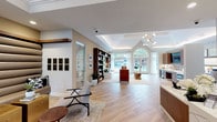 This elegantly designed single-floor community space blends contemporary style with warm, inviting details throughout. The grand entrance features tall glass-paneled white doors with arched transoms, flooding the foyer with natural light, while light-toned wood-style flooring flows seamlessly across the entire space.

The main lounge area is a standout, showcasing soaring tray ceilings with integrated ambient lighting and a striking wall of floor-to-ceiling walnut bookshelves. Generous arched windows dressed in sheer white drapery frame lush outdoor views, complementing a curated seating arrangement of leather chairs, a plush sofa, and eclectic accent tables.

An adjacent lounge nook features a dramatic horizontally channeled upholstered accent wall, a wall-mounted flat-screen TV beneath a floating wood shelf, and versatile seating — creating an intimate media and social area. The reception zone includes a sleek quartz-topped counter with rich wood cabinetry, a bold geometric tile backsplash, and a dedicated workstation area with natural light from large windows.

Throughout the space, a refined palette of warm beige, crisp white, and rich walnut tones creates a cohesive and welcoming atmosphere, accented by greenery and carefully curated décor that enhances the overall sense of polish and sophistication.
