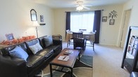 This single-story space showcases an open-concept layout that seamlessly connects the living and dining areas into one cohesive, functional space. The living area is anchored by plush carpeted flooring and features a comfortable seating arrangement centered around a rectangular coffee table, complemented by a decorative area rug underneath. A floor lamp and framed wall art add warmth and character to the space.

Adjacent to the living area, the dining space accommodates a round table with seating for multiple guests, positioned directly in front of a large window dressed with dark curtains. This window serves as a prominent architectural feature, allowing an abundance of natural light to brighten the entire open area and creating an inviting atmosphere throughout the day.

A ceiling fan with an integrated light fixture is centrally mounted overhead, providing both ventilation and additional ambient lighting. The neutral, light-toned walls complement the darker furnishings, creating a balanced contrast throughout the space. Overall, the layout offers a practical and comfortable environment suited for both everyday living and casual entertaining.