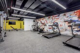 Located at 211 W Armour Blvd in Kansas City, Missouri, this fitness center is a well-equipped, single-floor facility designed to support a full range of workout routines. The space features an open layout with durable rubber flooring throughout, and an exposed black ceiling fitted with bright linear LED lighting that illuminates the entire room evenly.

A bold, large-scale abstract mural dominates one wall, adding a dynamic and energizing visual element to the environment. Complementing this, an accent wall painted in vivid yellow provides additional pops of color and visual energy, with a wall-mounted flat-screen TV positioned nearby for entertainment during workouts.

The cardio zone is well-appointed with treadmills, a stationary spin bike, and an elliptical machine, all arranged along the mural wall. The strength training area includes a Matrix cable machine system, a multi-functional rack, an adjustable weight bench, and a tiered dumbbell rack stocked with hex weights in a variety of sizes. A stability ball rounds out the functional training options.

A corridor connects additional areas of the facility, including what appears to be a secondary room or locker area with a bench for changing. The overall ambiance is modern, clean, and purposefully designed to motivate users, making this fitness center a comprehensive and functional amenity.