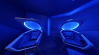 This single-floor wellness facility is defined by its striking, immersive atmosphere, where deep blue ambient lighting envelops every surface — from the walls and ceilings to the flooring — creating a calming, otherworldly environment. The centerpiece of the space is a collection of futuristic pod-style recliners, each featuring a clamshell-shaped canopy edged with glowing blue LED light strips. The pods are equipped with cushioned, ergonomically designed seating with integrated headrests, suggesting a focus on deep relaxation, meditation, or light therapy sessions. The pods are arranged to accommodate multiple users simultaneously, making the space suitable for group wellness sessions. Wall-mounted artwork depicting serene, atmospheric landscapes adds a contemplative visual element, reinforcing the tranquil ambiance. The flooring features a patterned carpet that complements the cool-toned lighting scheme. A transitional corridor area connects to the pod room, where neutral gray walls and standard lighting offer a subtle contrast to the vivid blue interior. Stainless steel accessory fixtures are present near the pod stations. The overall design of this space is purposeful and experiential, prioritizing sensory immersion and relaxation through deliberate use of color, light, and specialized equipment.