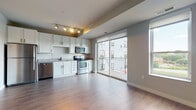 This single-floor residence showcases a contemporary open-concept design that seamlessly connects the kitchen and living area. The kitchen is well-appointed with white shaker-style cabinetry, granite countertops, a textured penny-tile backsplash, and a full suite of stainless steel appliances, including a refrigerator, electric range, over-the-range microwave, and dishwasher. Track lighting overhead highlights the kitchen's clean, modern aesthetic. Durable wood-look flooring extends throughout the main living space, complementing the neutral gray walls and creating a cohesive, contemporary feel.

The living area is bright and airy, benefiting from abundant natural light streaming through a large sliding glass door that opens directly onto a private balcony with a railing, offering outdoor views of the surrounding building. An additional side window further enhances the natural light in the space.

The bedroom is generously proportioned, featuring the same wood-look flooring, a sliding closet door, and access to a carpeted walk-in closet equipped with built-in wire shelving for ample storage. The bathroom includes a full-size soaking tub and shower combination with chrome fixtures, wood-look flooring, and clean white walls, providing a simple and functional design.