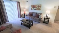 This single-floor residence offers a thoughtfully arranged living space designed for everyday comfort and functionality. The living room serves as the heart of the home, centered around a plush, neutral-toned sofa accented with decorative throw pillows in warm burgundy and soft teal hues. A dark-finished wood coffee table anchors the seating area, complemented by coordinating end tables and warm ambient lighting from table lamps. Soft wall-to-wall carpeting runs throughout the living area, adding warmth underfoot and contributing to the overall cozy atmosphere. A large sliding glass door fitted with vertical blinds and elegant drape panels floods the room with natural light while providing a seamless connection to the exterior. The neutral wall tones serve as a versatile backdrop, allowing the vibrant floral wall art to stand out as a visual focal point. A ceiling-mounted HVAC vent ensures year-round climate comfort. The overall design aesthetic is clean and inviting, with a balance of functional furnishings and decorative touches that create a lived-in, welcoming ambiance throughout the space.