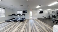 This single-level fitness center offers a thoughtfully organized workout environment divided into distinct functional zones. The space is finished with a neutral gray-and-white color palette, white crown molding, and a striped multi-tone carpet that runs throughout the primary cardio area, lending a clean, polished aesthetic.

The cardio zone is generously sized, housing multiple treadmills, upright stationary bikes, an elliptical, and a rowing-style machine. Multiple wall-mounted flat-screen televisions are positioned throughout the space for entertainment during workouts. Natural light enters through several large windows fitted with roller shades, offering views of surrounding greenery while allowing light control.

A dedicated strength and stretching zone features rubberized flooring, a weight bench, and a wall-mounted TV. This area is separated from the main cardio floor, providing a quieter space for focused training. An open shelving unit serves as a divider between the two zones.

Additional amenities include a medicine ball rack with an assortment of weighted balls, a water dispenser, a paper towel dispenser, and a mirrored wall section. A glass-paned exterior door provides convenient direct access to the outdoors. Overhead fluorescent lighting ensures the entire space remains well-illuminated throughout the day.