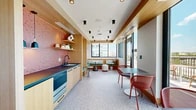 This compact yet highly functional space combines a fully equipped kitchen with a stylish dining and lounge area, all unified by a cohesive, contemporary design aesthetic. The kitchen is appointed with light maple cabinetry, a dark countertop, a stainless steel sink with a matte black faucet, and a built-in appliance drawer. A striking full-height backsplash of rose-pink hexagonal mosaic tiles serves as the kitchen's defining visual feature, complemented by a trio of pendant lights in matte black. Open wood shelving above displays decorative objects and greenery, adding warmth and personality.

The dining area flows seamlessly from the kitchen and is anchored by terrazzo flooring that runs throughout the entire space. Matte black cylindrical track lighting punctuates the wood-clad ceiling above. Built-in banquette seating with green cushions and patterned throw pillows occupies a nook partitioned by warm wood panels, with decorative circular wall art creating an artistic focal point. Small sculptural tables complete the lounge seating arrangement.

Floor-to-ceiling sliding glass doors and expansive windows flood the dining area with abundant natural light while framing sweeping city skyline views. The doors open directly onto a balcony, extending the usable space outdoors. Red curved dining chairs paired with dark tables provide additional seating near the windows, making this an inviting space for both casual gatherings and quiet moments.