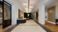 This thoughtfully designed single-floor space presents an impressive blend of modern sophistication and inviting warmth. The grand entry vestibule features sleek black-framed glass doors that flood the space with natural light, transitioning into a spacious lobby anchored by a curved, fluted dark wood reception desk. Behind the desk, a striking living moss wall bearing a prominent "H" monogram serves as a dramatic focal point, complemented by a textured stone accent wall and a plush navy velvet sofa with a curated seating arrangement.

Overhead, a linear track of pendant lighting runs the length of the corridor, drawing the eye through the space and highlighting its generous ceiling height and clean white arched crown moldings. The flooring transitions artfully between patterned mosaic tile at the entry and warm wood planks throughout the interior.

The café area is exceptionally well-appointed, featuring a marble-topped island, open black metal shelving, stainless steel appliances including a display case and draft taps, and globe pendant lighting. Adjacent lounge areas offer intimate dark-paneled booth seating with natural light from steel-framed windows, while a rounded dining room showcases lush green velvet banquette seating, marble bistro tables, and sheer-curtained windows — creating a refined yet comfortable gathering environment throughout.