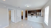This single-floor residence offers a thoughtfully designed open-concept layout that promotes a natural flow between living and kitchen areas. The kitchen is a standout feature, outfitted with rich dark-stained cabinetry that provides ample storage, light granite countertops, and stainless steel appliances including a refrigerator, microwave, and range. A breakfast bar peninsula extends from the kitchen, offering additional seating and serving as a functional divider between the kitchen and the adjoining living space. Recessed lighting and flush-mount ceiling fixtures keep the space well-lit throughout. Large, polished light-toned tile flooring runs continuously across the open areas, adding a sense of cleanliness and spaciousness. The soft gray-white wall tones complement the overall neutral palette, creating a bright and cohesive atmosphere. A hallway off the main living area leads to additional rooms, including what appears to be a bathroom with vanity lighting visible in the background. Louvered bi-fold closet doors and a standard panel door are also visible near the hallway entry, suggesting practical storage solutions. The overall design is clean, modern, and move-in ready.
