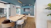 This conference room is thoughtfully designed to balance professional functionality with contemporary style. The space is immediately distinguished by its teal-painted ceiling, which adds a bold visual contrast against the white walls and creates a refined, modern atmosphere. A large marble-topped conference table serves as the room's centerpiece, surrounded by a mix of curved upholstered chairs and wood-accented seating that provide both comfort and elegance. Suspended above the table, a sleek brass linear pendant light adds a warm, sophisticated glow that complements the recessed ceiling lighting.

A wall-mounted flat-screen display is positioned on the teal accent wall, making the space well-suited for presentations and collaborative meetings. Textured carpet underfoot adds warmth and acoustic softness to the room. Natural light filters in from nearby windows, contributing to a bright and open feel.

One of the room's standout features is its glass railing along one side, which opens visually to a multi-level common area below, complete with hardwood flooring, a credenza with decorative artwork, and an adjacent lounge. This transparency creates a sense of connectivity while maintaining a dedicated, purposeful meeting environment.