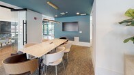 This conference room is thoughtfully designed to balance professional functionality with contemporary style. The space is immediately distinguished by its teal-painted ceiling, which adds a bold visual contrast against the white walls and creates a refined, modern atmosphere. A large marble-topped conference table serves as the room's centerpiece, surrounded by a mix of curved upholstered chairs and wood-accented seating that provide both comfort and elegance. Suspended above the table, a sleek brass linear pendant light adds a warm, sophisticated glow that complements the recessed ceiling lighting.

A wall-mounted flat-screen display is positioned on the teal accent wall, making the space well-suited for presentations and collaborative meetings. Textured carpet underfoot adds warmth and acoustic softness to the room. Natural light filters in from nearby windows, contributing to a bright and open feel.

One of the room's standout features is its glass railing along one side, which opens visually to a multi-level common area below, complete with hardwood flooring, a credenza with decorative artwork, and an adjacent lounge. This transparency creates a sense of connectivity while maintaining a dedicated, purposeful meeting environment.