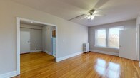 This single-floor unit offers a warm and functional layout highlighted by beautifully polished hardwood floors that run throughout the main living areas and bedrooms, reflecting natural light and lending a sense of warmth to every room. The living area is spacious and open, featuring a ceiling fan with light fixture, a cast-iron radiator, and a window that brings in natural light with views of the surrounding neighborhood. A closet area with an exposed rod provides convenient storage directly off the main living space. The kitchen is efficiently laid out with warm-toned wooden cabinetry, granite-look countertops, a gas range, and a full-size refrigerator, offering all the essentials in a compact footprint. The bedroom is generously proportioned with its own ceiling fan, radiator, and large windows that frame outdoor views, creating a bright and comfortable retreat. A second room or flex space connects seamlessly to the layout and offers additional flexibility. The bathroom is a standout feature, showcasing a classic clawfoot soaking tub with an overhead shower, crisp white wainscoting, a vanity with mirror, and a timeless black-and-white tile floor. The entry foyer provides a practical transition space with a clothing rod. Throughout, neutral gray walls and white trim create a clean, cohesive aesthetic.