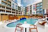Located at 3100 Lake Villa Dr in Metairie, Louisiana, this outdoor amenity area is a standout feature of a modern, multi-story residential community. The centerpiece is a generously sized swimming pool with a freeform design, highlighted by striking blue-tiled waterfall features that cascade dramatically into the main pool. A separate, circular spa pool with its own water jets offers a more intimate relaxation option alongside the main swimming area. The pool deck is finished in light-toned tile and is surrounded by an abundance of cushioned chaise lounges, teak-accented benches, and shaded seating areas with large market umbrellas, providing both sun and shade options for residents. Warm wood-slat privacy fencing encloses the space, creating a sense of seclusion while complementing the contemporary architectural palette of the surrounding building. The multi-story residential structure frames the courtyard on multiple sides, incorporating balconies, large windows, and a mix of white, gray, and red facade panels. A covered outdoor pavilion area with an upper-level overlook terrace adds architectural depth. Accent palm trees and stylish furnishings throughout contribute to the resort-inspired ambiance of this well-appointed outdoor retreat.