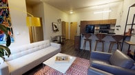 This single-floor space showcases a thoughtfully designed open-concept layout that seamlessly blends the living and kitchen areas into one cohesive environment. The living area is anchored by a tufted light grey sofa paired with a sleek, glass-topped coffee table resting on a bold red and white chevron area rug. A striking yellow arc floor lamp adds a pop of color and warmth, complementing the eclectic wall décor that includes framed artwork and sculptural accents. Dark hardwood-style flooring runs throughout, providing a rich contrast to the neutral wall tones. A small bistro table and wire chairs tucked near the entryway offer an intimate dining nook. The kitchen features warm walnut-toned cabinetry, a dark countertop breakfast bar, and modern appliances including a microwave and refrigerator. Two sculpted mid-century modern bar stools provide casual seating at the bar. The overall ambiance is modern and curated, blending functional design with personality-driven décor elements that create an inviting and stylish living environment.