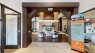 This single-floor community clubhouse or leasing center is a thoughtfully designed space that blends contemporary style with warm, rustic accents. The grand entrance features floor-to-ceiling glass doors that flood the foyer with natural light, complemented by large-format white tile flooring that transitions into rich wood-look flooring throughout the interior.

The main lounge area commands attention with dramatically vaulted ceilings adorned with exposed wood beams and a striking chandelier. A sleek linear fireplace set against a floor-to-ceiling stone tile surround serves as the room's focal point, flanked by neutral-toned lounge chairs arranged for conversation. Bold teal accents punctuate the space through furnishings and architectural details, lending a vibrant, modern energy.

Decorative laser-cut metal wall panels add an artful layer of texture, while reclaimed wood columns ground the open layout with organic warmth. A game and entertainment area features a billiards table and additional seating nooks with upholstered banquette benches and curated wall art.

The kitchenette is outfitted with stone countertops, a hexagonal tile backsplash, undermount sink, and appliances. A separate meeting or leasing room offers a coffered ceiling, teal accent walls, exposed brick, a round conference table, and views to the outdoors through large windows.
