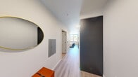 Located at 513 1st Ave N in Seattle's Lower Queen Anne neighborhood, this efficiently designed one-bedroom, one-bathroom unit offers a thoughtful layout that maximizes every square foot. Light wood-tone flooring runs throughout, creating a warm, cohesive aesthetic across the entire space.

The entry hallway sets a clean, modern tone with crisp white walls, a recessed utility panel, and a round gold-framed mirror that adds a stylish accent. The hallway leads naturally into the main living area, where an open-plan kitchen and sleeping space share a generously sized room bathed in natural light from large windows overlooking Seattle's cityscape and surrounding greenery.

The kitchen is outfitted with stainless steel appliances — including a range, microwave, and refrigerator — complemented by light wood upper cabinetry, dark lower cabinets, and a subway tile backsplash. The bedroom area features a sliding barn door closet with matte black hardware, adding an industrial-modern touch to the otherwise light and airy space.

The bathroom is well-appointed with a dark espresso wood vanity topped with a white countertop, a large wall mirror with bright overhead lighting, and consistent light wood flooring that ties the space together. Clean lines and neutral tones throughout give this unit a polished, contemporary feel.