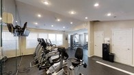 This fitness center is a single-floor facility designed to support a comprehensive workout experience. The space is divided into distinct workout zones, beginning with a cardio area outfitted with multiple elliptical trainers and a stationary bike, all positioned near a row of windows fitted with horizontal blinds that allow natural light to fill the room. The light-colored walls and recessed ceiling lighting create a bright, energizing atmosphere throughout.

Beyond the cardio zone, the space opens into a strength training area featuring a multi-function cable and weight machine, along with a free-weight bench and additional equipment. The dark rubber flooring throughout provides both safety and durability, ideal for high-traffic exercise environments. The ceiling is smooth and finished with evenly spaced recessed lights, ensuring consistent illumination across the entire space.

A white door near the back offers access to an adjacent room or utility space, and a small mounted display near the entryway may serve as a directory or informational panel. The overall layout is open and functional, with thoughtful zoning that allows multiple users to exercise simultaneously without crowding. The clean, neutral aesthetic and well-maintained equipment make this a practical and welcoming fitness facility.