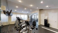 This fitness center is a single-floor facility designed to support a comprehensive workout experience. The space is divided into distinct workout zones, beginning with a cardio area outfitted with multiple elliptical trainers and a stationary bike, all positioned near a row of windows fitted with horizontal blinds that allow natural light to fill the room. The light-colored walls and recessed ceiling lighting create a bright, energizing atmosphere throughout.

Beyond the cardio zone, the space opens into a strength training area featuring a multi-function cable and weight machine, along with a free-weight bench and additional equipment. The dark rubber flooring throughout provides both safety and durability, ideal for high-traffic exercise environments. The ceiling is smooth and finished with evenly spaced recessed lights, ensuring consistent illumination across the entire space.

A white door near the back offers access to an adjacent room or utility space, and a small mounted display near the entryway may serve as a directory or informational panel. The overall layout is open and functional, with thoughtful zoning that allows multiple users to exercise simultaneously without crowding. The clean, neutral aesthetic and well-maintained equipment make this a practical and welcoming fitness facility.