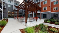 This outdoor courtyard is a thoughtfully designed amenity space nestled within a modern multi-story residential community. The building exterior features a contemporary combination of gray and warm orange horizontal lap siding, accented by black metal railings on the private balconies that overlook the courtyard below.

The courtyard is organized into several distinct functional zones. A central fire pit area is constructed with a stone base and surrounded by modern outdoor sofas and lounge chairs upholstered in bold orange, providing a welcoming gathering spot. Adjacent to this lounge zone, a substantial wood pergola with rich stained timber beams defines an al fresco dining area, furnished with multiple round tables and aluminum chairs arranged for group seating.

Connecting the various zones is a series of wide concrete walkways bordered by carefully maintained landscaping. Ornamental grasses, low shrubs, and fresh mulched planting beds line the pathways, adding greenery and texture throughout the space. A small lawn area is also visible, further softening the hardscape.

Ground-level units face directly onto the courtyard, with sliding glass doors and private patio areas separated by black metal railings and compact planting strips. Large windows allow views into interior common areas from the courtyard, reinforcing a sense of community connectivity. The overall ambiance is clean, modern, and inviting, offering residents a versatile outdoor environment for relaxing and socializing.