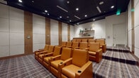 This dedicated media room delivers a refined, theater-style experience through a carefully considered design. Rows of plush, cognac-colored leather recliner seats are arranged in a tiered layout facing a large wall-mounted display screen, each seat equipped with built-in cup holders for added convenience. The dark, paneled ceiling with recessed lighting creates an immersive, cinema-like ambiance, while the patterned, multi-toned carpet grounds the space with a contemporary aesthetic.

The walls feature alternating acoustic panels and warm wood-toned vertical accents, contributing both to the visual appeal and sound quality of the room. A clerestory window above the screen wall allows a measure of natural light without disrupting the viewing experience.

Along the rear of the room, a well-appointed wet bar provides a practical amenity for entertaining. It includes a stone-topped counter, wood cabinetry, a built-in sink, stainless steel appliances, and floating wood shelves above, styled with decorative accessories. Together, these elements make the space well-suited for private screenings, presentations, or social gatherings in a polished, functional environment.