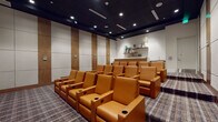 This dedicated media room delivers a refined, theater-style experience through a carefully considered design. Rows of plush, cognac-colored leather recliner seats are arranged in a tiered layout facing a large wall-mounted display screen, each seat equipped with built-in cup holders for added convenience. The dark, paneled ceiling with recessed lighting creates an immersive, cinema-like ambiance, while the patterned, multi-toned carpet grounds the space with a contemporary aesthetic.

The walls feature alternating acoustic panels and warm wood-toned vertical accents, contributing both to the visual appeal and sound quality of the room. A clerestory window above the screen wall allows a measure of natural light without disrupting the viewing experience.

Along the rear of the room, a well-appointed wet bar provides a practical amenity for entertaining. It includes a stone-topped counter, wood cabinetry, a built-in sink, stainless steel appliances, and floating wood shelves above, styled with decorative accessories. Together, these elements make the space well-suited for private screenings, presentations, or social gatherings in a polished, functional environment.